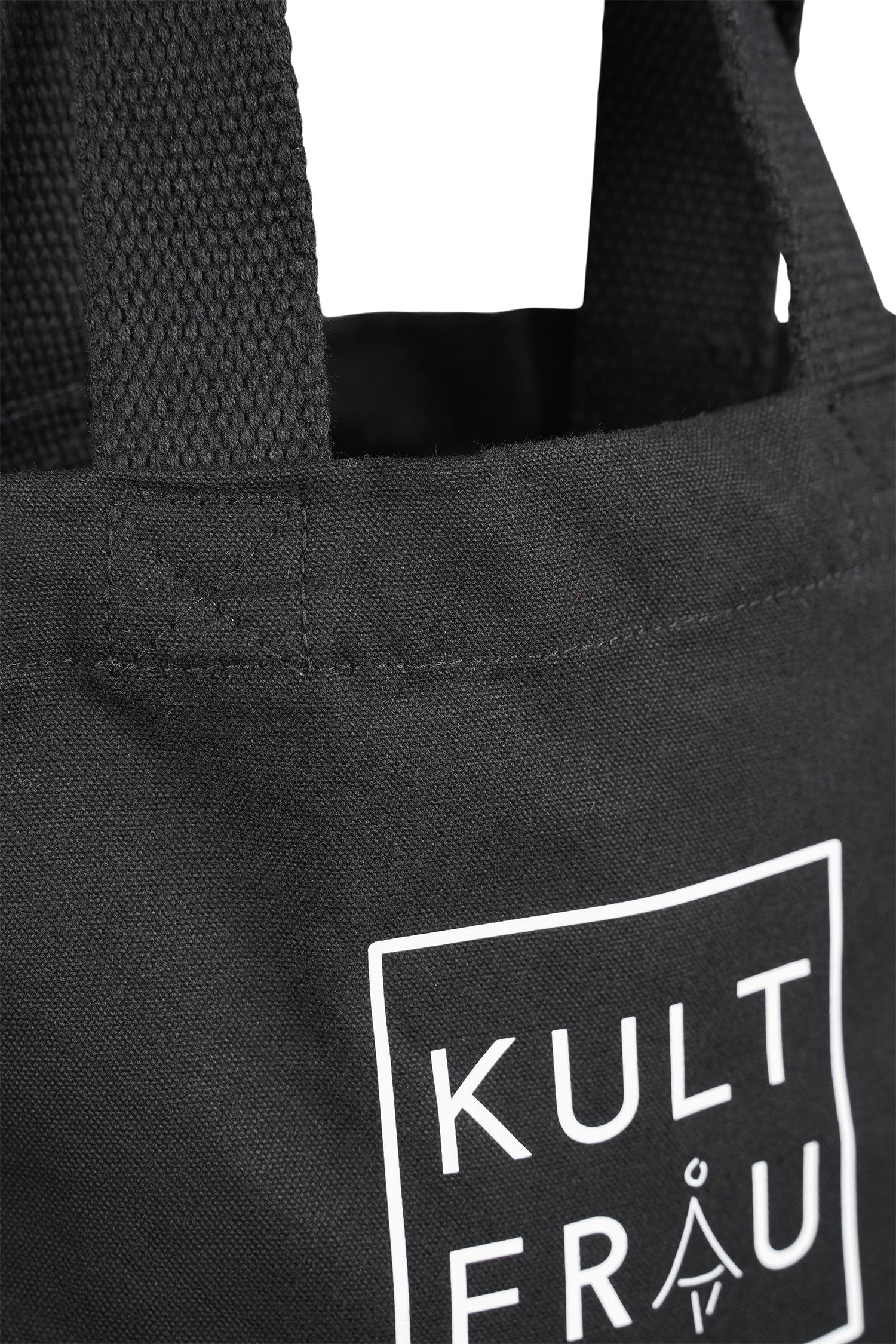 Nahaufnahme einer schwarzen Stofftasche aus robustem Material. Die strukturierte Oberfläche der Tasche ist gut sichtbar, ebenso wie ein Teil des weißen Logos auf der Vorderseite, das den Brandnamen "KULTFRAU" zeigt. Der kurze, stabile Henkel besteht aus gewebtem Stoff und ist mit einer verstärkten Naht in Kreuzform an der Tasche befestigt. Die hochwertige Verarbeitung der Nähte und des Materials ist deutlich erkennbar.