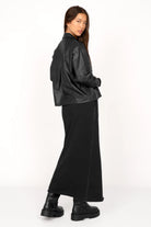 Woman wearing a cropped black Hemdjacke aus supersoftem Eco-Leder with Kent collar and chest pockets, styled with a long black skirt and boots.