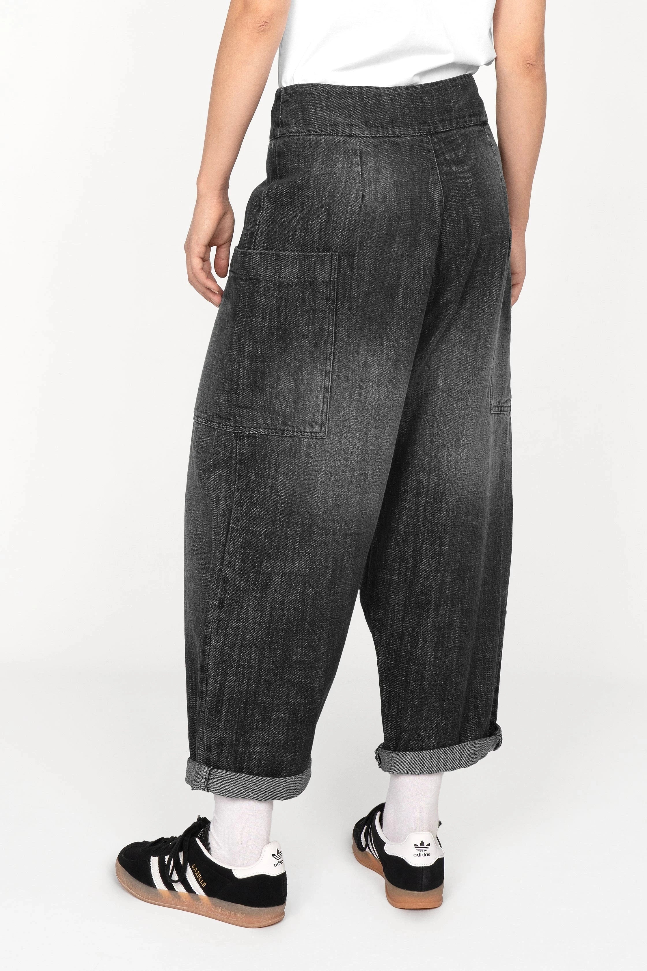 Back view of VIKADA Revive Baggy Hose in black gradient wash with deep crotch, large side pockets, button fly, and rolled cuffs, worn with white socks and black sneakers.
