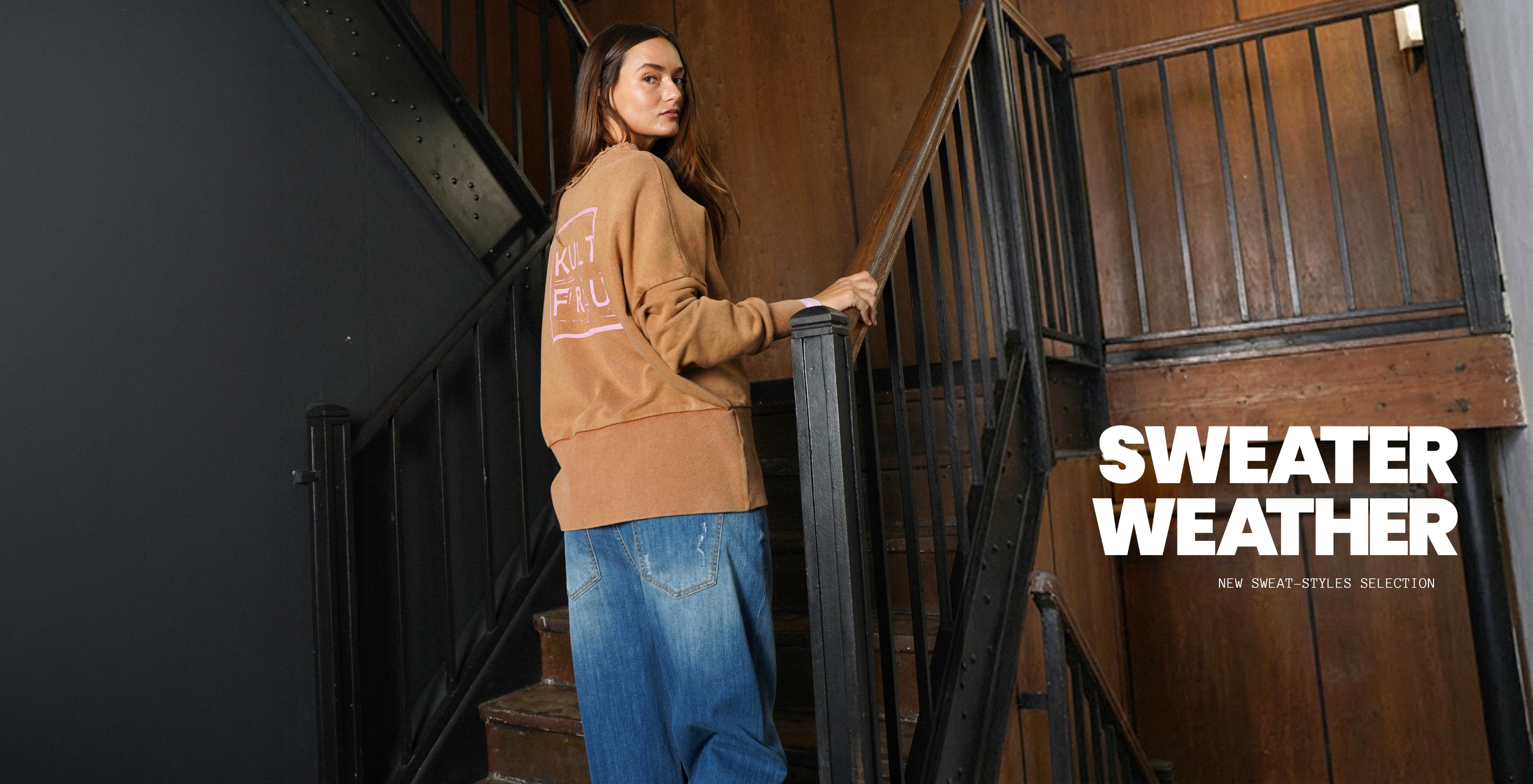 Banner mit Model auf einer Holztreppe, trägt braunes KULTFRAU-Sweatshirt und blaue Jeans; rechts steht der Text ‚SWEATER WEATHER‘ und ‚New Sweat-Styles Selection‘.