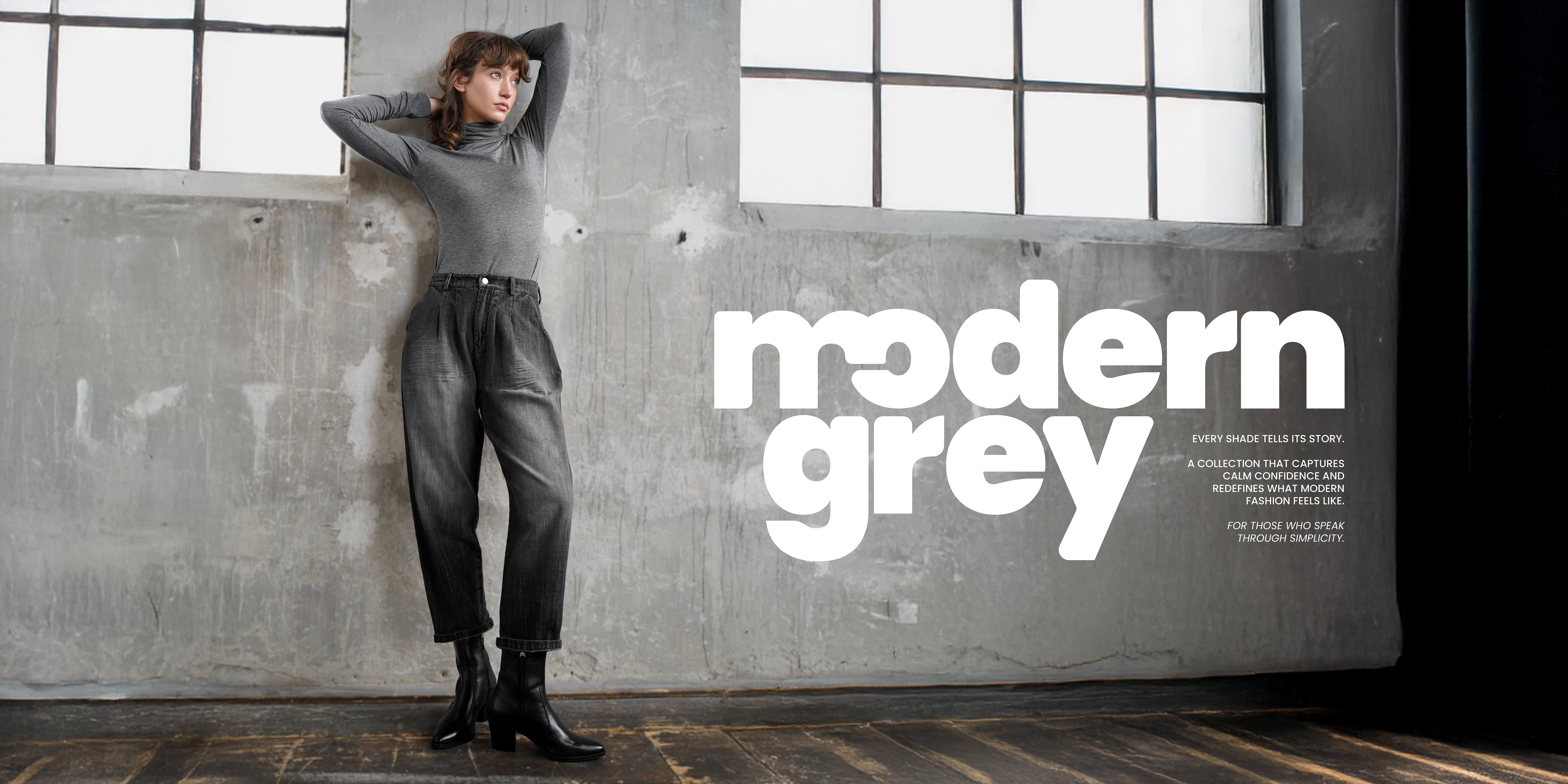 Frau trägt einen grauen Rollkragenpullover und eine verwaschene graue Jeans mit Bundfalten, kombiniert mit schwarzen Ankle Boots. Im Hintergrund Betonwand mit der Aufschrift ‚modern grey‘ – minimalistischer, urbaner Look in Grautönen.